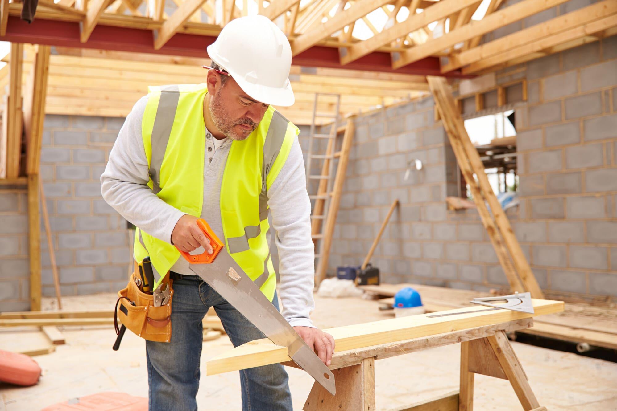 Carpenter sawing timber on a sawhorse at an outdoor construction site with roof frame behind