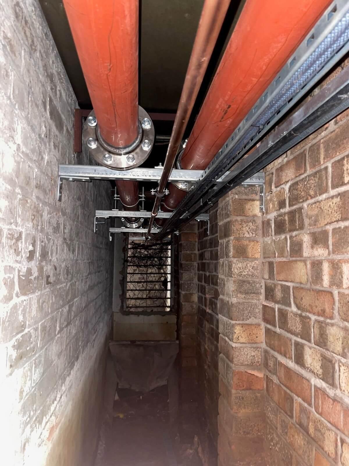 Industrial riser duct with large red and copper pipes running along brick walls