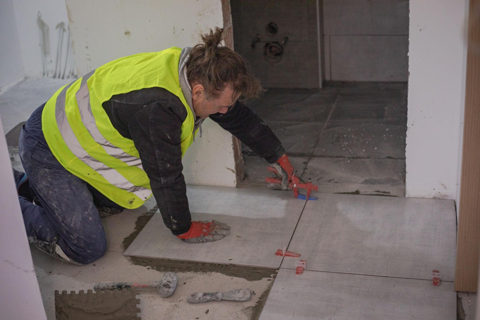 Tiler in hi-vis kneeling and levelling floor tiles with spacers at a wall junction
