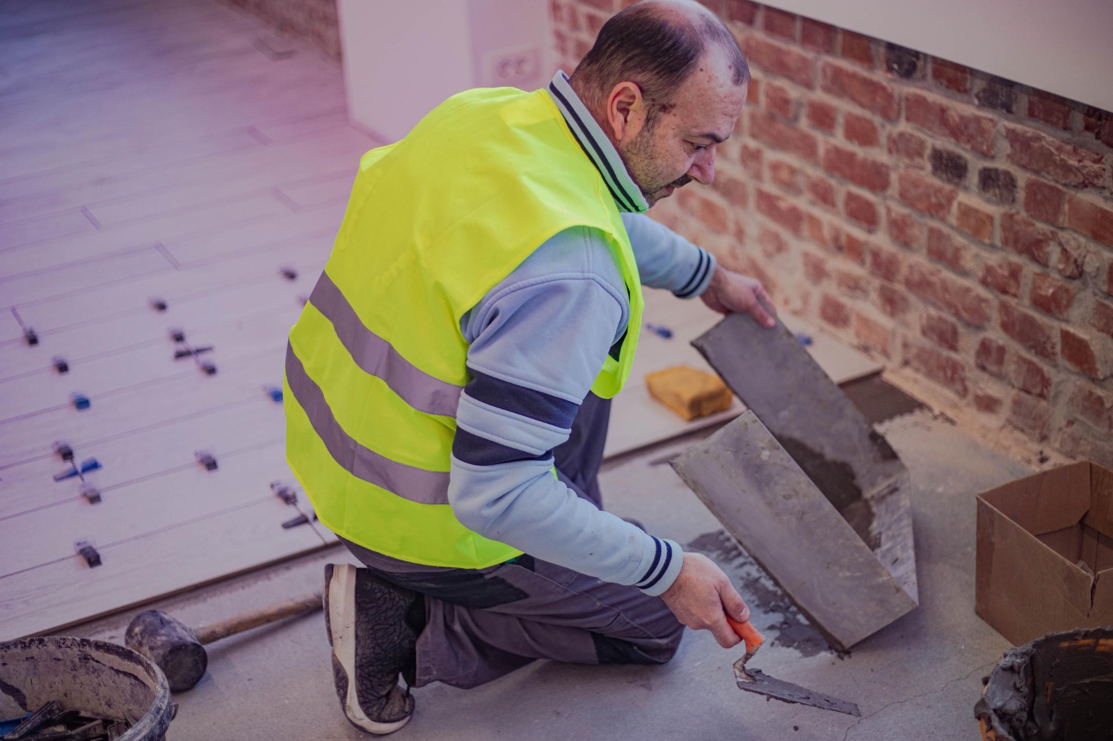 Tiler in hi-vis applying adhesive and setting floor tiles on an under-construction floor